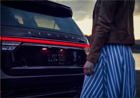 A person is shown near the rear of a 2023 Lincoln Aviator® SUV as the Lincoln Embrace illuminates the rear lights | Seekins Lincoln in Fairbanks AK