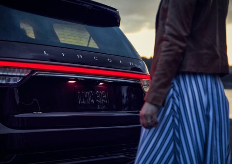 A person is shown near the rear of a 2024 Lincoln Aviator® SUV as the Lincoln Embrace illuminates the rear lights | Seekins Lincoln in Fairbanks AK
